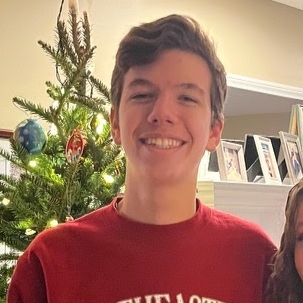 A young man with short hair smiles at the camera in front of a Christmas tree.