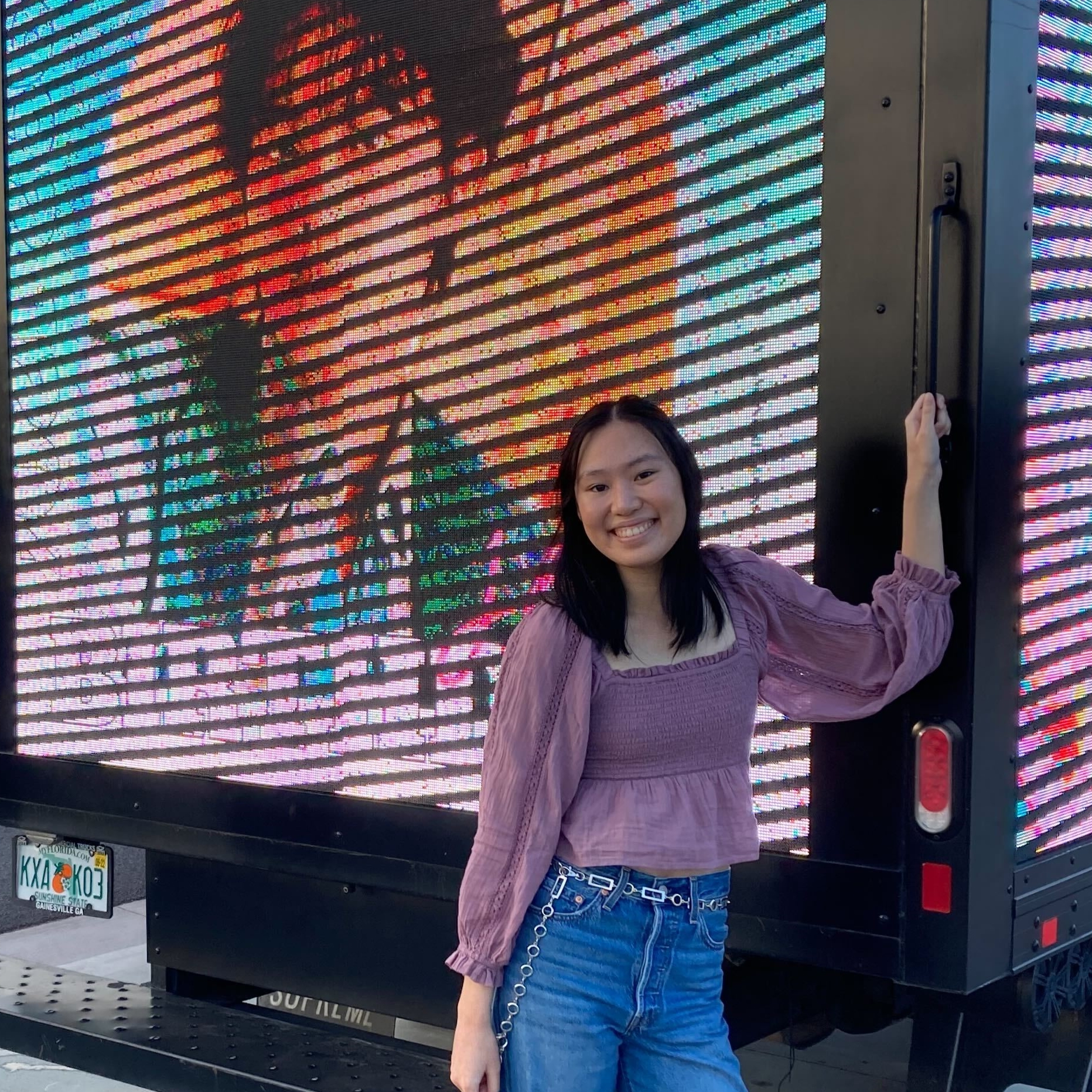 A young Asian woman smiles at the camera in front of a truck with a celebrity painted on it.